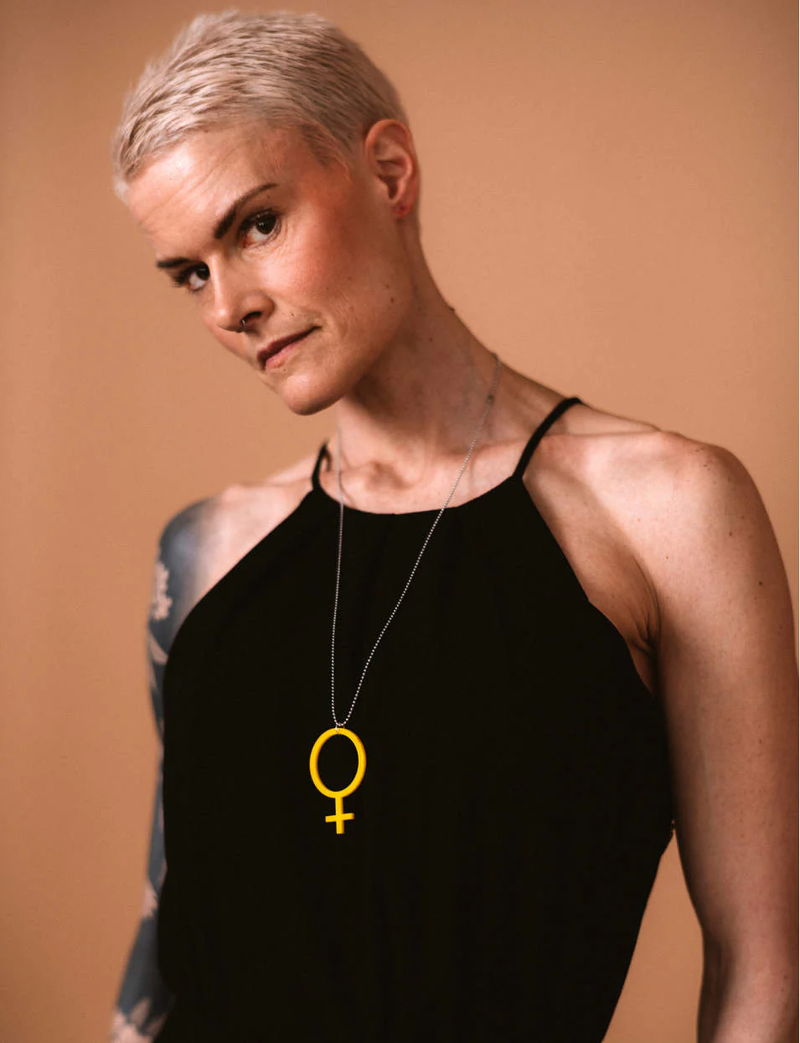 Kvinna med kort blont hår och svart linne med smala axelband. Hon bär ett halsband med en stor gul feminist-symbol och står mot en beige bakgrund.