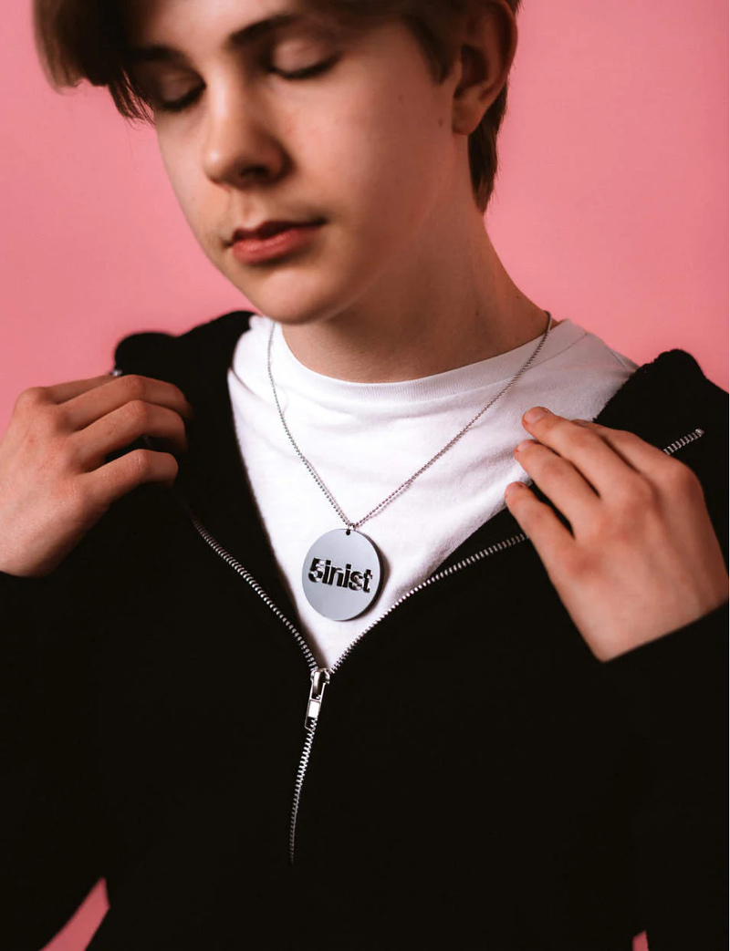 Ung man med kort ljust hår, vit T-tröja och svart hoodie. Han bär ett runt halsband i metall med utskuret ordet “feminist” och står mot en rosa bakgrund.